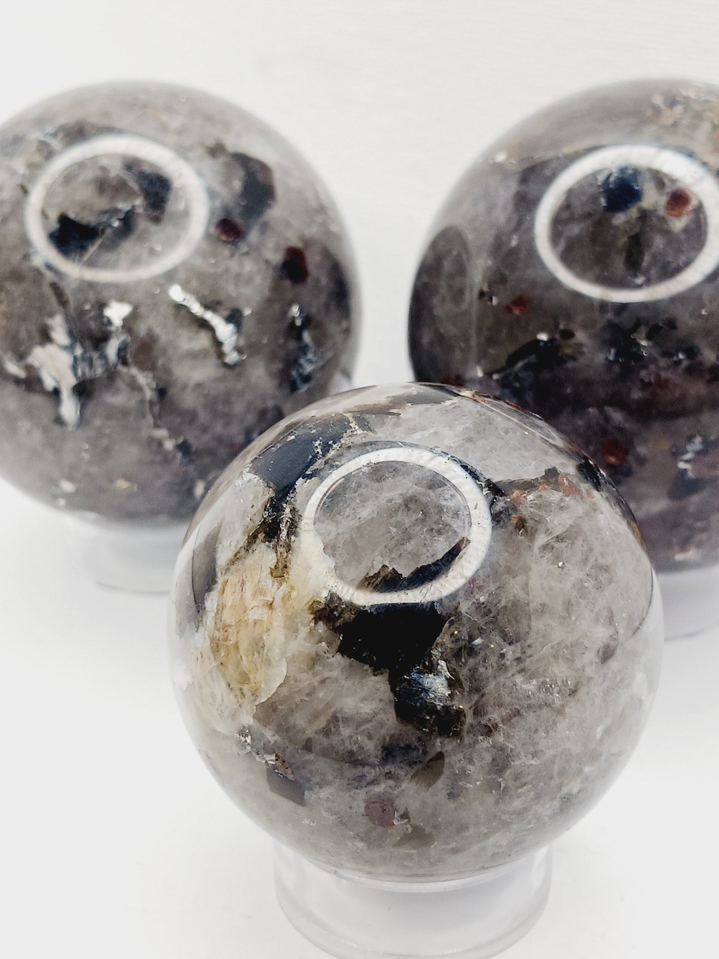 Quartz, garnet, tourmaline, and mica sphere, polished with clear, red, black, and shimmering inclusions, displayed on a white background.