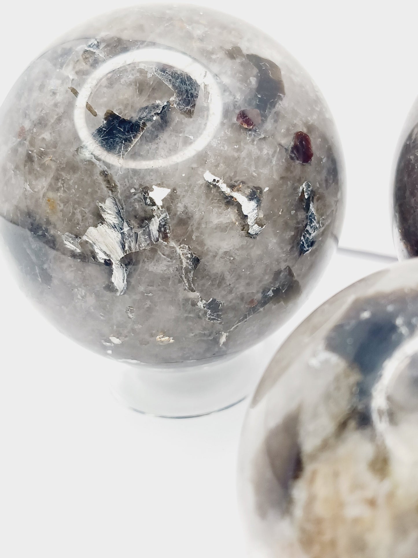 Quartz, garnet, tourmaline, and mica sphere, polished with clear, red, black, and shimmering inclusions, displayed on a white background.