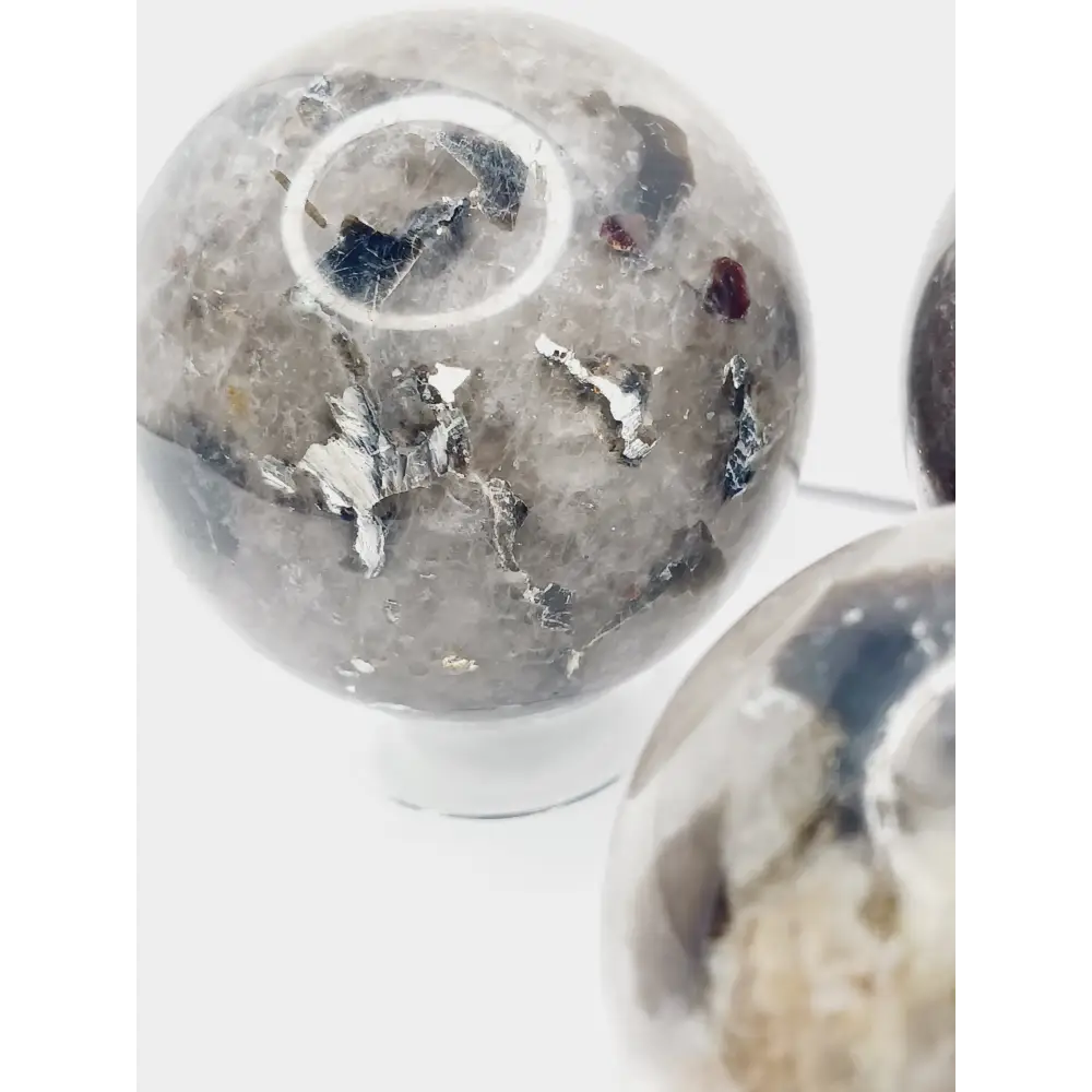Quartz, garnet, tourmaline, and mica sphere, polished with clear, red, black, and shimmering inclusions, displayed on a white background.