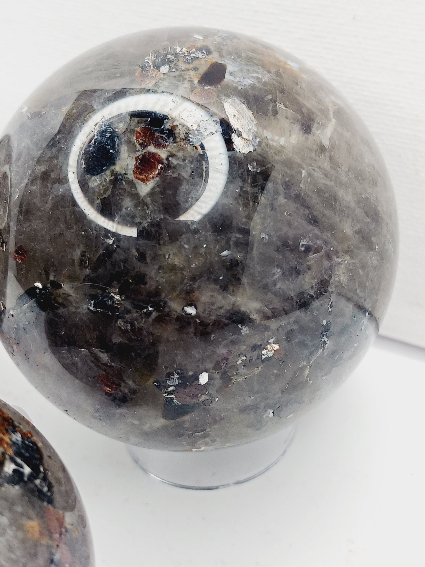 Quartz, garnet, tourmaline, and mica sphere, polished with clear, red, black, and shimmering inclusions, displayed on a white background.