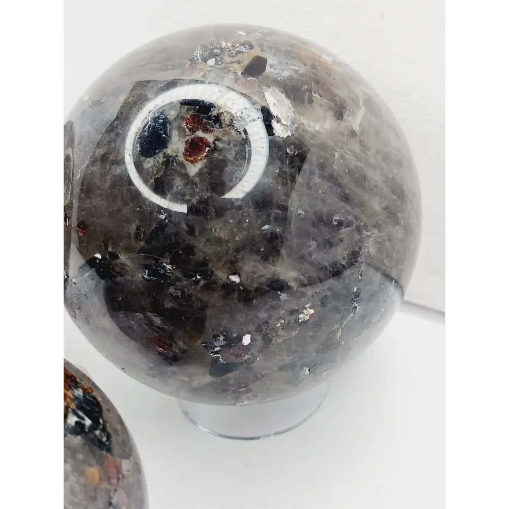 Quartz, garnet, tourmaline, and mica sphere, polished with clear, red, black, and shimmering inclusions, displayed on a white background.