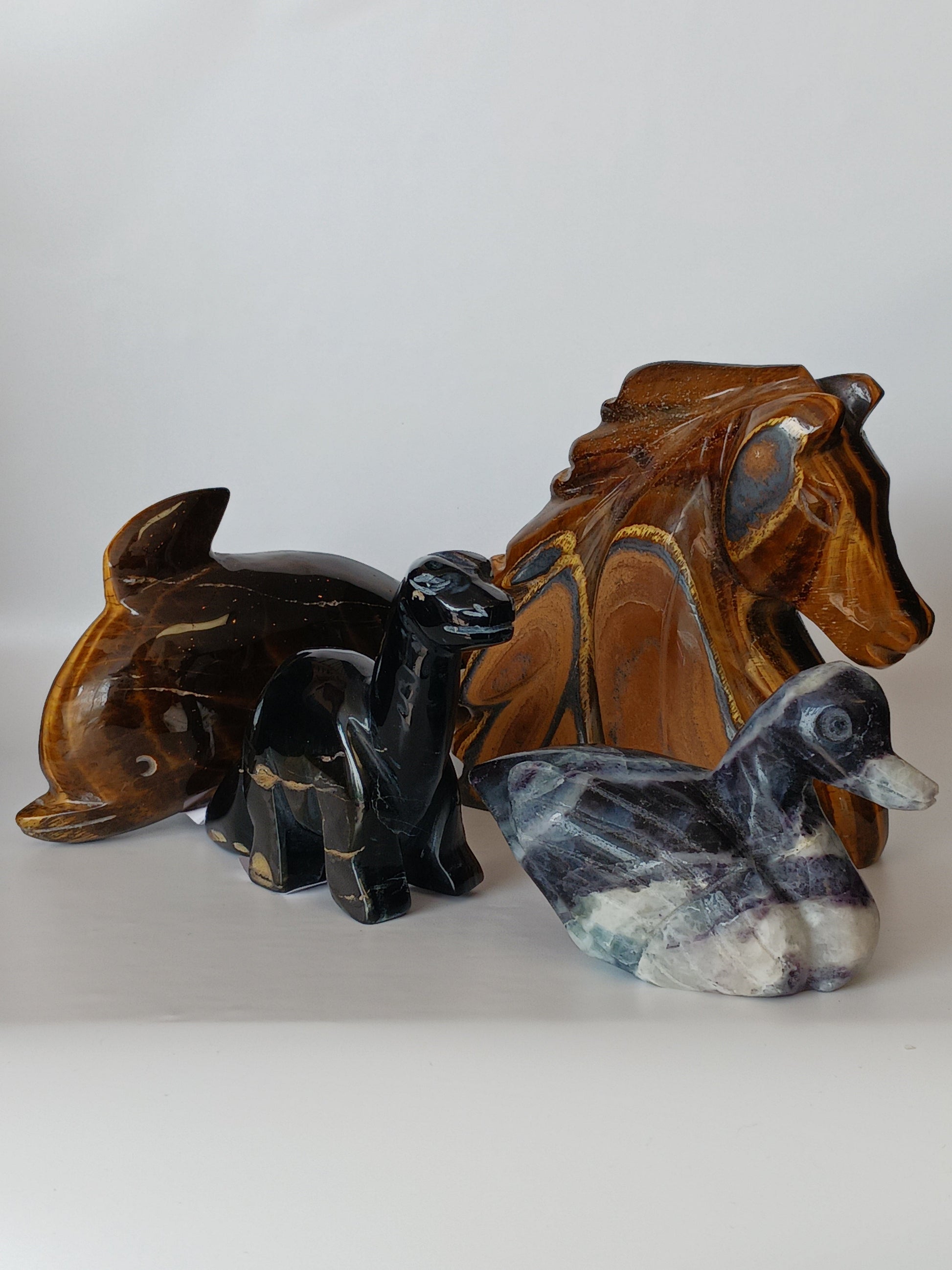Assorted hand-carved animal figurines made from Blue Tiger’s Eye, Fluorite, and Tiger’s Eye crystals, each featuring unique colors and patterns, displayed on a white background.