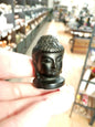 Hand-carved 1 1/2 inch tall Buddha head figurines in assorted crystal materials such as amethyst, rose quartz, clear quartz, and jade, displayed on a white background.