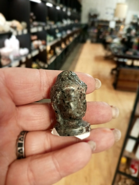 Hand-carved 1 1/2 inch tall Buddha head figurines in assorted crystal materials such as amethyst, rose quartz, clear quartz, and jade, displayed on a white background.