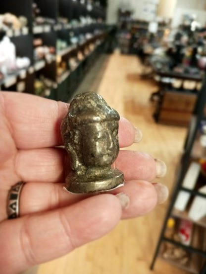 Hand-carved 1 1/2 inch tall Buddha head figurines in assorted crystal materials such as amethyst, rose quartz, clear quartz, and jade, displayed on a white background.
