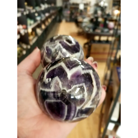 Hand-polished Dream Amethyst sphere featuring vivid purple and white chevron bands, displayed on a white background.
