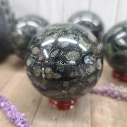 Hand-polished Kambaba Jasper sphere featuring deep green and black swirling orbicular patterns, displayed on a white background.