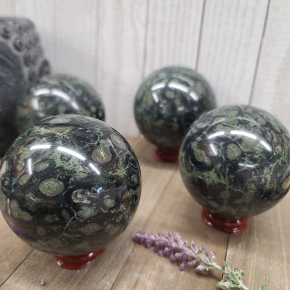 Hand-polished Kambaba Jasper sphere featuring deep green and black swirling orbicular patterns, displayed on a white background.