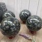 Hand-polished Kambaba Jasper sphere featuring deep green and black swirling orbicular patterns, displayed on a white background.