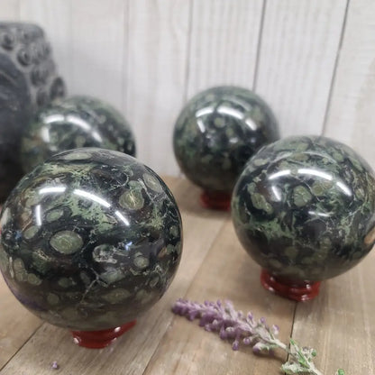 Hand-polished Kambaba Jasper sphere featuring deep green and black swirling orbicular patterns, displayed on a white background.