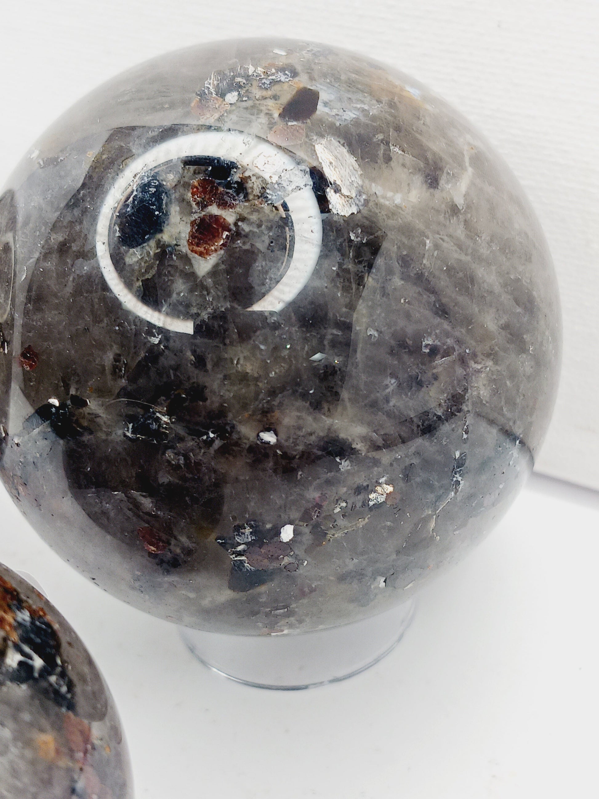 Quartz, garnet, tourmaline, and mica sphere, polished with clear, red, black, and shimmering inclusions, displayed on a white background.