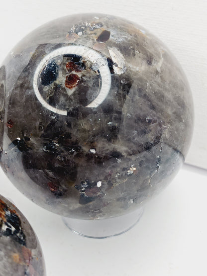Quartz, garnet, tourmaline, and mica sphere, polished with clear, red, black, and shimmering inclusions, displayed on a white background.