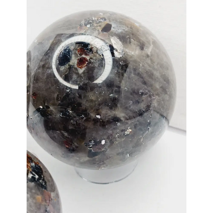 Quartz, garnet, tourmaline, and mica sphere, polished with clear, red, black, and shimmering inclusions, displayed on a white background.