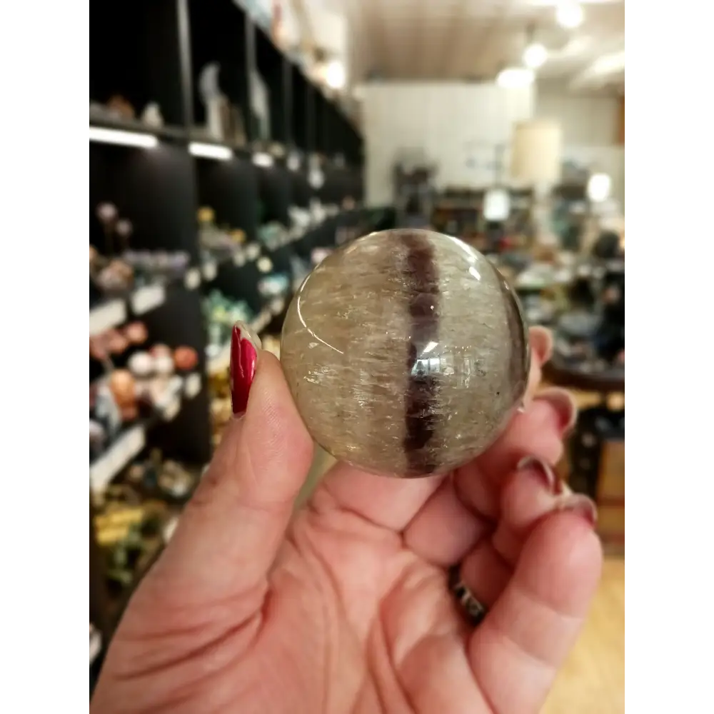 Hand-polished Rainbow and Clear Fluorite sphere featuring luminous bands of purple, green, blue, and clear tones, displayed on a white background.
