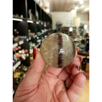 Hand-polished Rainbow and Clear Fluorite sphere featuring luminous bands of purple, green, blue, and clear tones, displayed on a white background.
