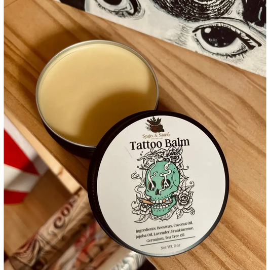 Tattoo Healing Balm in a small jar, surrounded by fresh herbs and natural oils, displayed on a white background. Perfect for soothing, protecting, and revitalizing tattoos.

